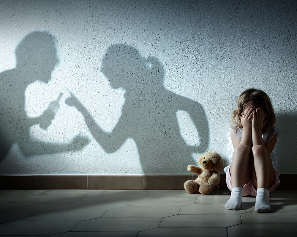 A child sits on the floor with here hands over her face while her parents shadows on the wall are fighting with each other representing a child experiencing trauma who could benefit from Trauma Counseling in Birmingham, AL.