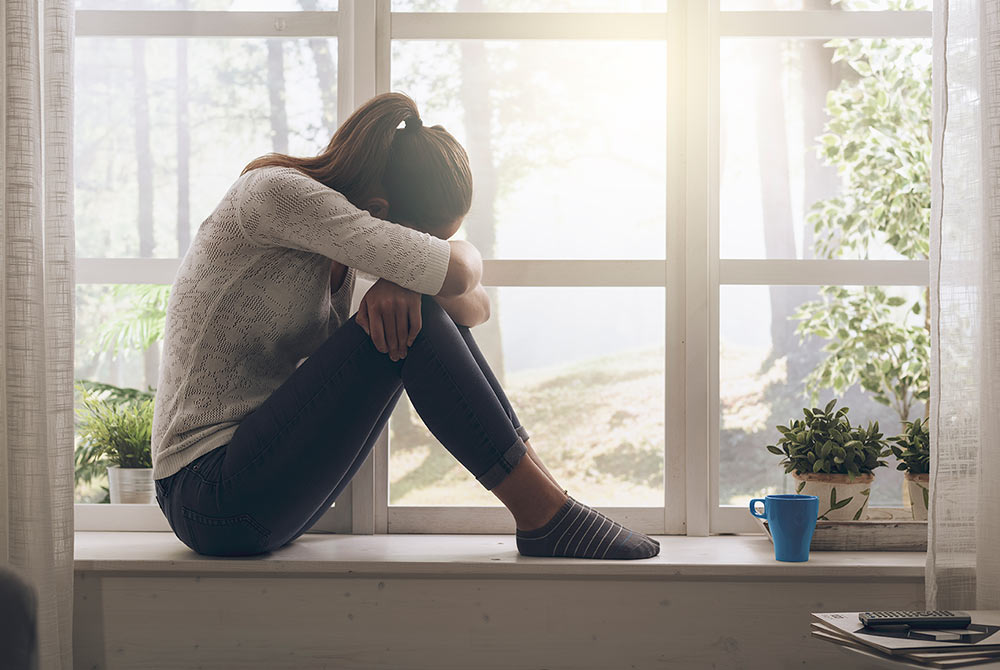 A woman sits on a window sill with her head on her knees as she thinks about her depression symptoms. She's decided to seek depression treatment with Empower Counseling in Birmingham, AL 35223.