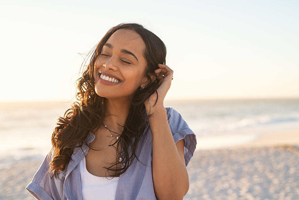 Image of a smiling woman tucking her hair behind her ear while standing on the beach with her eyes closed. Discover effective ways to manage your anxiety from trauma with EMDR therapy in Birmingham, AL.