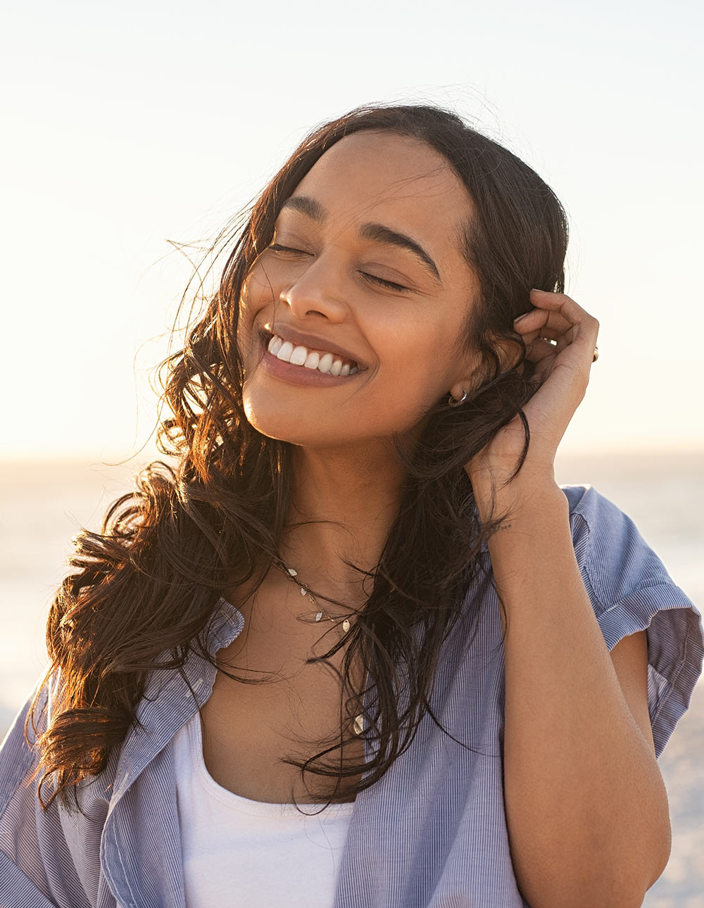 Image of a smiling woman closing her eyes and tucking her hair behind her ear while standing on the beach. Uncover a positive way to manage your trauma with EMDR therapy in Birmingham, AL.